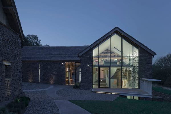 A stone 16th century, now a modern day barn conversion, photo taken at dusk showing a two storey glazed wall, through the glass can be seen a floating staircase with a curving steel banister and a a seating area on the first floor mezzaine.