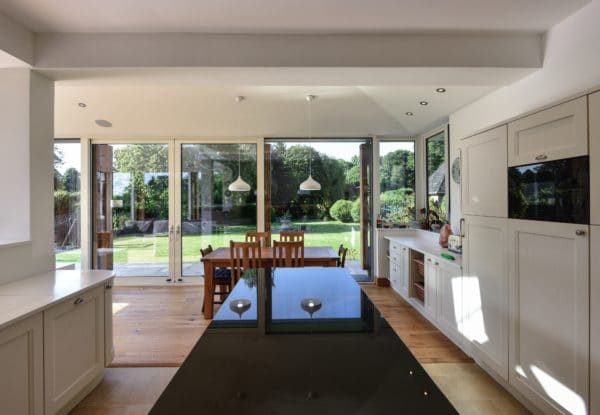 Interior of Home Extension. A black, marble kitchen island in a white kitchen, in the background the wooden dining table can be seen with a glazed wall behind it. Communion Architects Herefordshire.