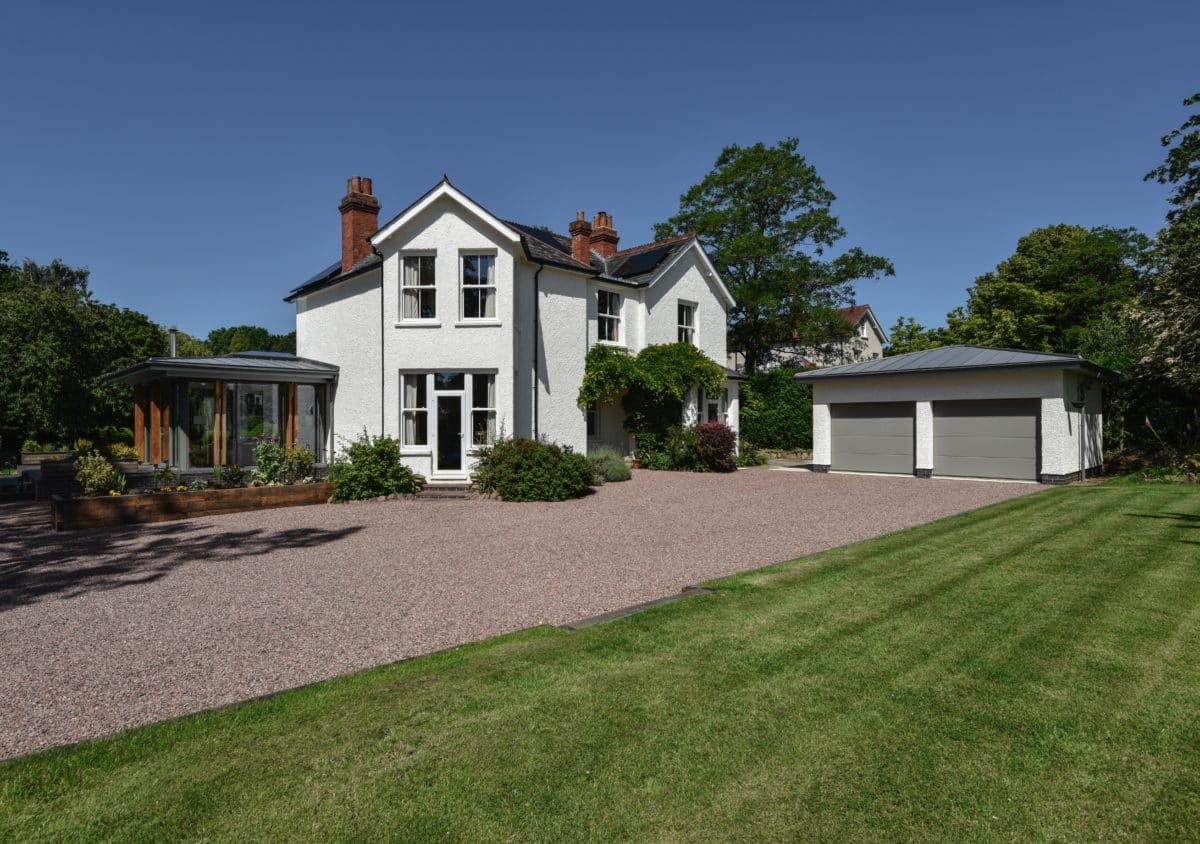 A stone driveway leading to a double garage and white 1930s house. To the left hand side of the house is a Home extension by Communion Architects.