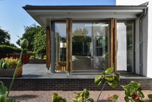 Side view of home extension by communion architects, Herefordshire. Image shows flat, over hanging roof projecting full height glazing, framed with aluminium and timber columns on a raised patio.