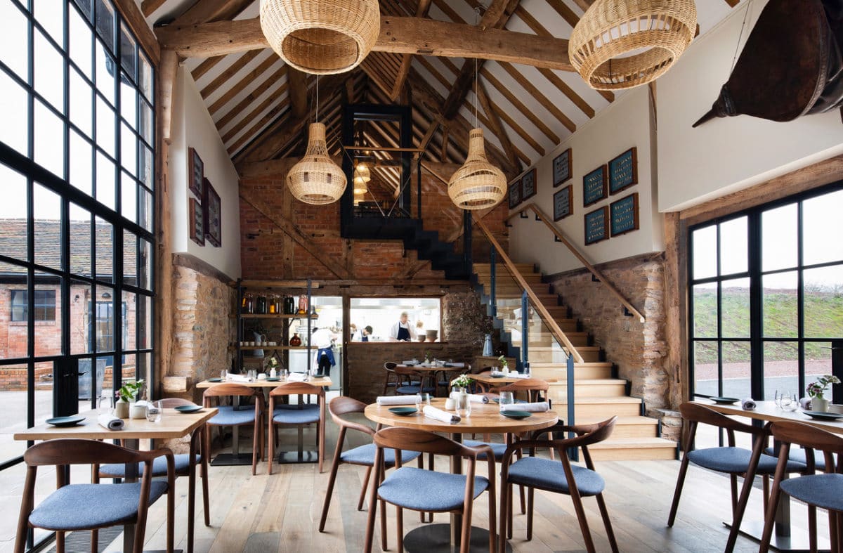 Interior of Pensons Restaurant on the Herefordshire/Worcestershire border. This was a derelict 16th century barn which has been transformed into a light-filed Michelin starred restaurant by Communion Architects. Two large steel-framed windows are opposite each other, between them is a timber floor with several wooden tables and chairs. Down the the length of the former barn is the open kitchen with chefs busy cooking. At the end on the right hand side is a floating staircase leading to a first floor private dining room. The walls have exposed stone and the rafters are painted white with exposed timber beams. From these hang four handwoven wicker lampshades. Communion Architects, Barn Conversion, Herefordshire.