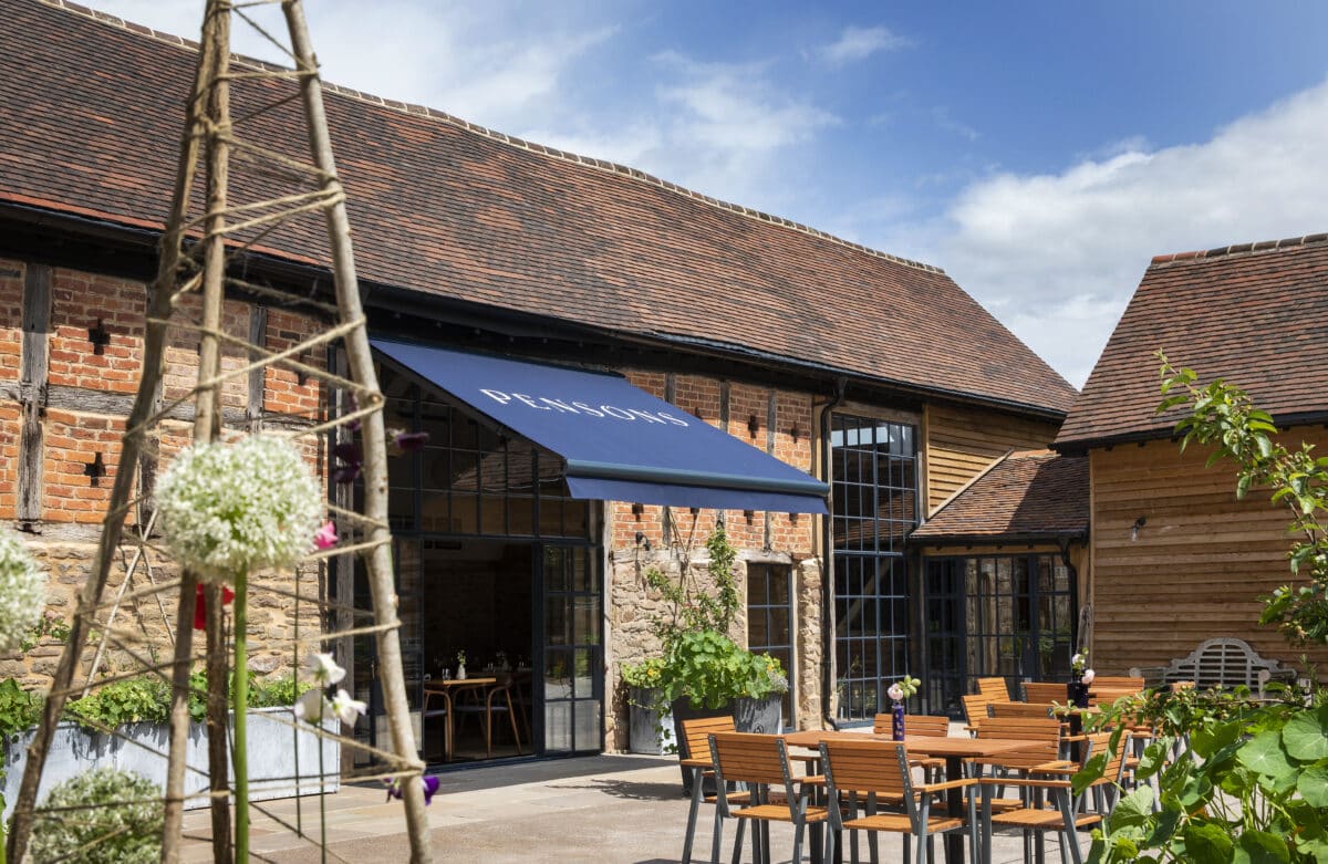 Exterior of Pensons restaurant, in the foreground is seating in the courtyard area, in the background is the restaurant, a 16th century barn with stone foundations, timber supports, red brick and a clay tiled roof.