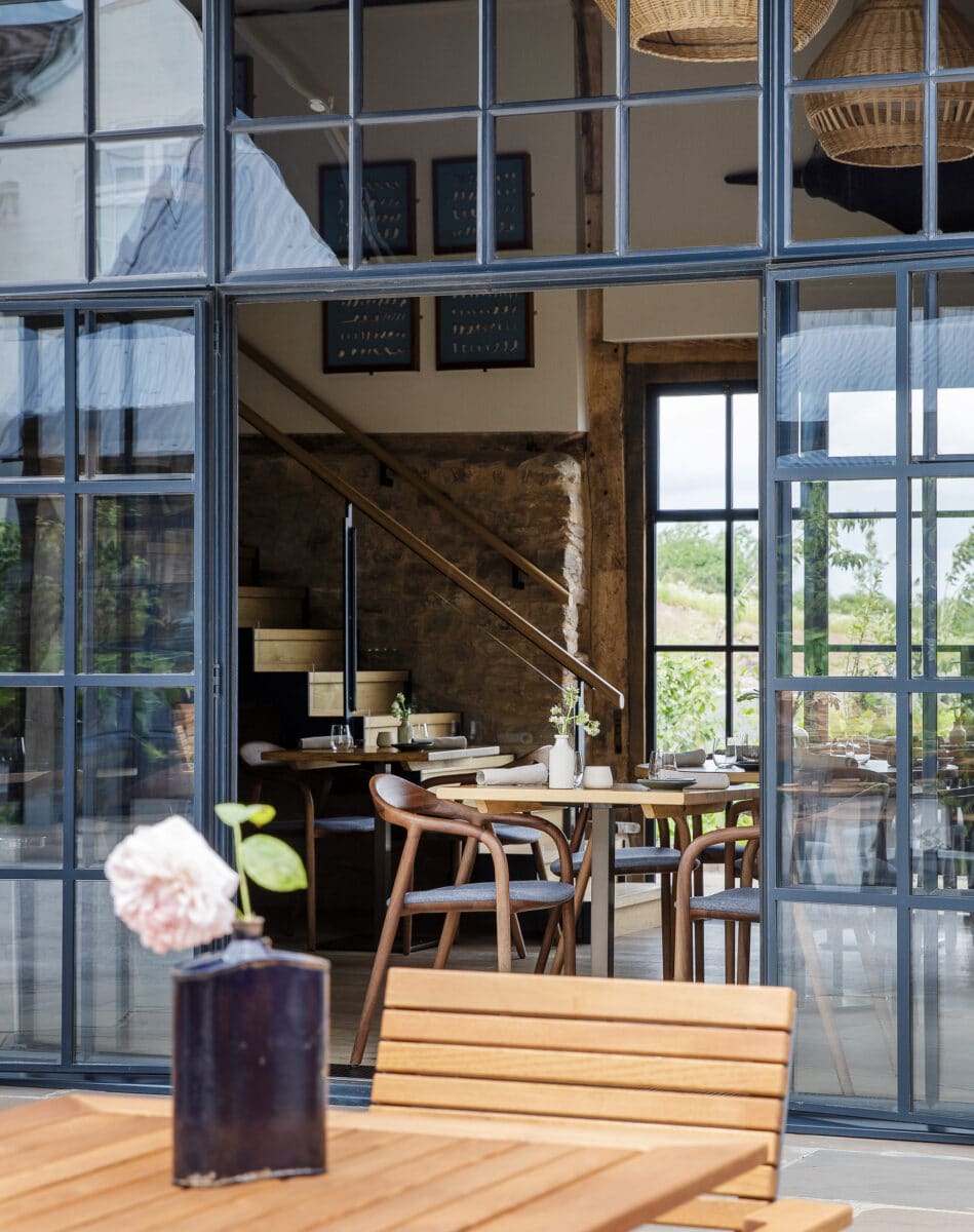 Pensons restaurant, barn conversion. Photo taken from outside, looking through steel-framed windows into the restaurant., inside are modern wooden chairs and tables with a sense of rustic charm.