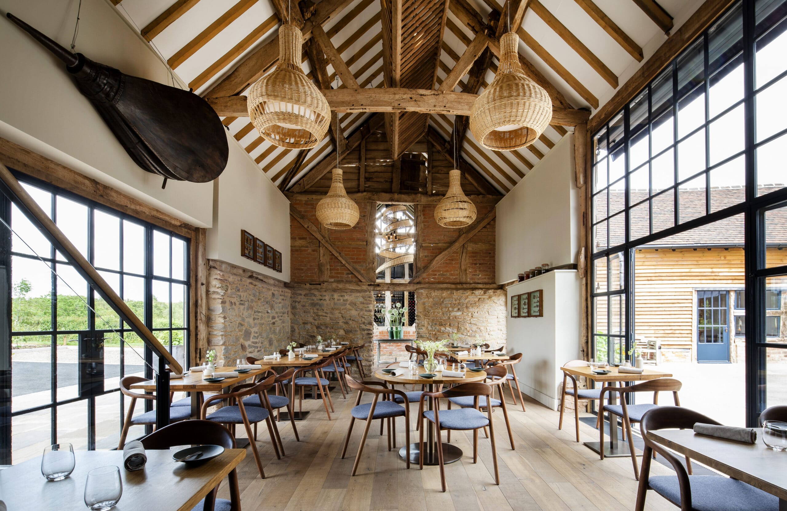 Interior of Pensons Restaurant, a barn conversion. Light-filled area interior from steel-framed windows on either side, above area exposed timber trusses with large bespoke wicker lampshades. Inside is a timber floor with seating. A modern interior with a sense of rustic charm.