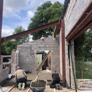 Constructing an extension. A red steel frame in the foreground and a breeze block wall in the foreground, in-between are two builders laying a screed floor.