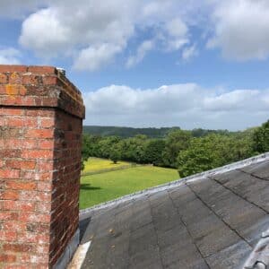A view from the roof. A slate roof and read brick chimney in the foreground with a green field and woodland in the background. Working on a home extension to a listed Georgian building in Herefordshire. Communion Architects.