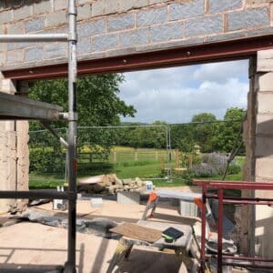 On a construction site, a breeze brick wall with a large opening in it which will form a window, through the opening can be see green fields and a woods in the background.