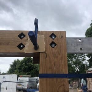 Image from construction site. The top right corner of an empty timber door frame is visible. The horizontal and vertical pieces are being temporarily held together with a joinery tools while they are secured. Through the top lintel runs a steel barn, providing extra support. Communion Architects, Herefordshire.