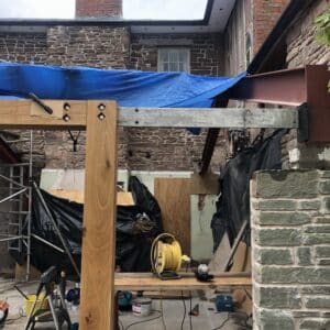 On the site of listed building in Herefordshire - constructing an extension. Image shows a low stone wall with an empty doorway frame next to it. A steel beam runs through the top timber beam proving extra support. The stone listed building can be seen in the background, communion architects Herefordshire