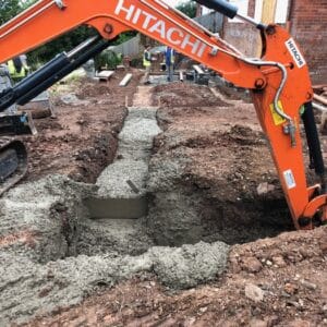 A bright orange arm of digger ploughs into the soil to create a trench for cement foundations. Communion Architects, Herefordshire