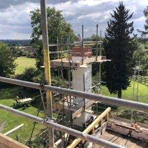 Looking across out over green field, evergreens and a cloudy blue sky from the roof of a fire damaged cottage, the view is interrupted in the foreground by the scaffolding and chimney breast being repaired. Communion Architects, Herefordshire