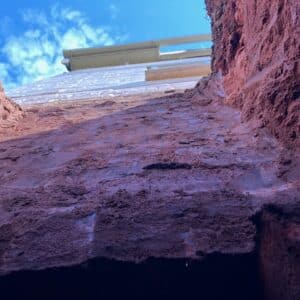 Standing inside some deep foundations looking up to the clear blue sky. The soil is a beautiful dark red reflecting the Herefordshire location, above it rises the existing house with the sky above.