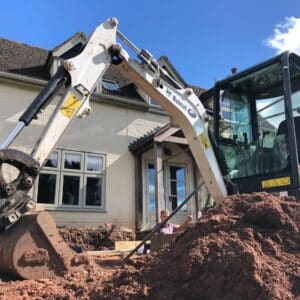A small digger with a white arm on site. The bucket rests on the floor behind the pile of earth in the foreground, a two storey White cottage in the background. On Site for an extension in Worcestershire, communion architects.