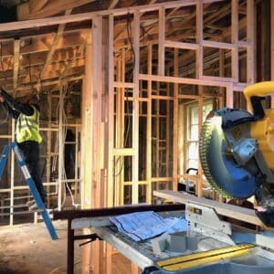 A builder in a high viz jacket in the back ground surrounded by a timber frame structure he is creating for an extension, a circular saw sits in the foreground. Communion Architects, Hereford.