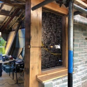 A new oak window frame set in a newly built stone wall, through the empty window a builder in a hi-viz jacket and white hard hat can be seen surveying works. Communion Architects, Hereford.