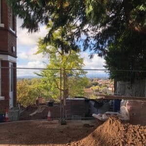On the left hand side is a red brick victorian house with large windows, on the far right side is dark green foliage, in the centre, there is a cityscape view across Herefordshire partially blocked by a green tree in the centre. Communion Architects, Hereford.