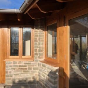 Exterior of a domestic dwelling, L-shaped extension with stone walls and oak door and window frames, protected by a low overhanging roof. Communion Architects, Herefordshire.