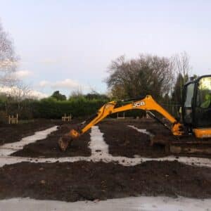 A small yellow digger with an extended arm reaching across a muddy ground. The ground is mostly mud with some poured concrete lines in a rectangular formulation, constituting the beginning of some foundations for a new house. Communion Architects, Hereford.