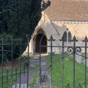 A wrought iron gate with one side slightly open. Behind the gate is a grassy graveyard and stoney path leading to the main door of a village church behind. The stone walls are a light yellow stone and the roof is pale brown. The church is partially obstructed by a large evergreen. Communion Architects, Hereford.