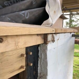 Close up of roof detailing a new oak beam connected with wooden pegs to an older wooden post which in turn is connected to a white washed stone wall. Communion Architects, Hereford.