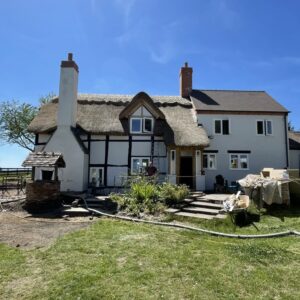 A black and white timber framed house with a thatched roof and large chimney breast on the left hand side. The right hand side has a double height extension with small windows, white render and tile roof, with grass in front. Communion Architects, Hereford, Herefordshire.
