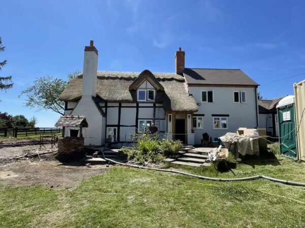 A black and white timber framed house with a thatched roof and large chimney breast on the left hand side. The right hand side has a double height extension with small windows, white render and tile roof, with grass in front. Communion Architects, Hereford, Herefordshire.