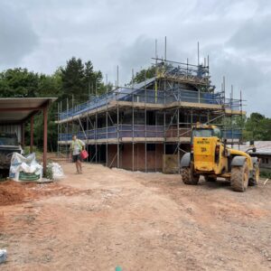 a muddy building site with Herefordshire's red clay soil. In the middle of the dirt is a scaffold-covered bungalow, a new dwelling. A yellow digger rests in front of the new house under construction. Communion Architects, Hereford, Herefordshire.