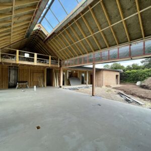 Interior of a new dwelling in Herefordshire, under construction. Concrete floor, timber beams, exposed insulation, in the pitched roof. The pitched roof has a pitched window running the length of the peak. The walls show exposed breeze blocks and brick, with the right hand wall open for floor to ceiling windows. Communion Architects Hereford.