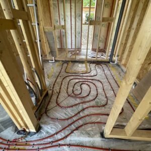 Timber framed wall building, part way through construction, with no packing between the timber frames yet. On the floor is a curling red tube curling across the floor expanse - part of an underfloor heating system. communion Architects Hereford, Herefordshire.
