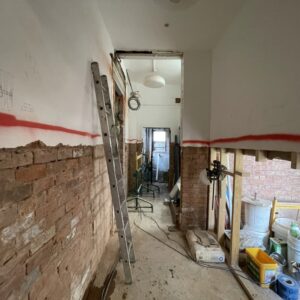 A corridor strewn with building equipment such as tool boxes and ladders. The top half of the walls are painted white, the lower half have been stripped and show exposed brick. A red line runs between the two. Communion Architects Hereford.