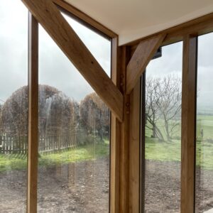 A portrait image of a corner of a room where two floor to ceiling windows meet with oak beam framing and bracing the space. cream roof and garden outside with a miuddy flower bed, grass and trees beyond. Communion Architects, Hereford, Herefordshire.