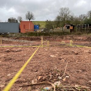 A muddy field, red Herefordshire clay soil, with yellow rope marking out rectangular zones. Preparing to dig foundations. Communion Architects, Hereford, Herefordshire.