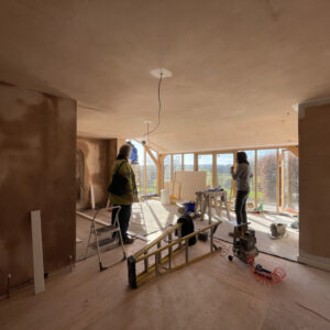 A freshly plastered room, light brown, still slightly damp. Two people stand in the space looking at the room and out the end window, the far wall has floor to ceiling glass windows looking out to a green grassy view. A new extension by communion architects, Hereford, Herefordshire.