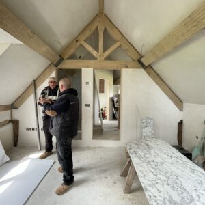 Pitched roof attic room with two men dress in black surveying the work done. Timber beams and Alex supports are visible. Plaster boards scatter the floor for use in another room. Communion architects, Hereford, Herefordshire.