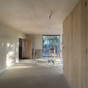 Looking through an oblong room, freshly plastered walls, timber cupboard doors to one side and full height glazed windows at the far end. Communion Architects, Hereford.