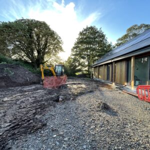 A small yellow digger clearing a muddy area while in the right hand side of the image is a new single storey house with a slate roof and glass and timber walls. Communion Architects Hereford.
