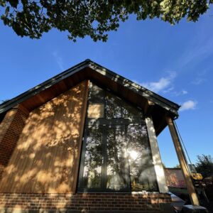 end facade of a new house designed by communion architects - one storey with an apex roof, with foundations of brick supporting one half of a glazed wall and half of a timber covered facade.