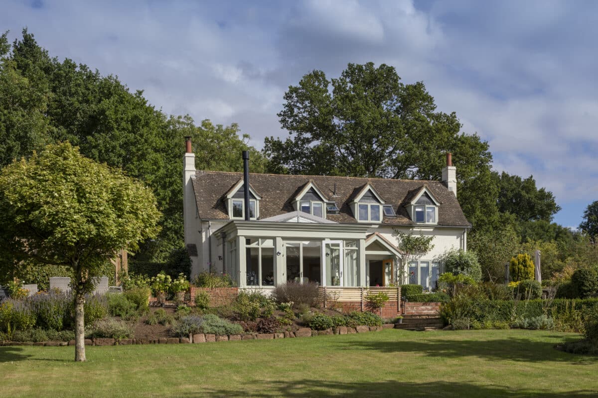 A pitched roof cottage set in a green and leafy garden surroundings. On the left of the cottage is a delicate timber and glazing extension. Sensitively added and in keeping with the cottage character of the house. Communion Architects, Hereford.